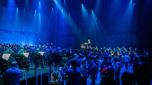 A large audience stands in the middle of a concert hall surrounded by a full orchestra performing on three sides. The conductor is elevated on a central platform, facing the musicians and leading the performance. Blue stage lighting creates a dramatic, immersive atmosphere, and beams of light shine down from above. The scene blends classical music with a modern, interactive experience as the audience is positioned within the orchestra, rather than in traditional seating.