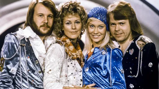 This is a photo of the pop group ABBA. They are standing closely together in a line, two on the left facing the other two on the right, like they are hugging, Benny on the far left, Anni-Frid next to him, next to her Agnetha Fältskog and Björn Ulvaeus far right. Benny and Anni-Frid have their right shoulders and faces to the camera, and Agnetha and Björn have their left shoulders towards the camera.