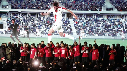 This is a photo of the Georgian National Men's Football standing out a darkened stage with fans and media below them filming and taking photos. There is a screen showing a football game as the backdrop of the stage.