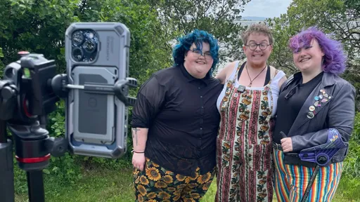 Three smiling people pose together outdoors in front of greenery and water, while a smartphone on a tripod records them. The person on the left has short teal hair, glasses, and wears a black sheer shirt with sunflower-print pants. The middle person has short hair, glasses, and wears floral-striped overalls over a white top. The person on the right has bright purple hair and wears a black jacket with colorful pins, a black shirt, and rainbow-striped pants. All three appear happy and relaxed in a natural setting.