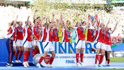 A jubilant women's football team in red and white Arsenal kits celebrates on a stage with confetti falling around them. One player holds the UEFA Women's Champions League trophy aloft, while others cheer and raise their fists in victory. The stage backdrop reads "WINNERS" and "UEFA Women's Champions League Final," with the Arsenal logo visible. All players wear gold medals around their necks.