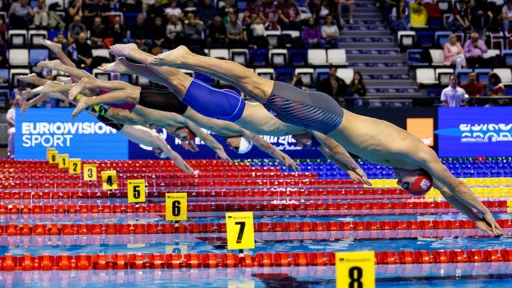 A group of male swimmers mid-dive at the start of a competitive race in an indoor swimming pool. Each swimmer is captured in perfect form above their respective lanes, which are marked with numbered yellow lane markers from 1 to 8. The pool is lined with red lane dividers, and the background shows spectators in the stands and a "Eurovision Sport" banner.