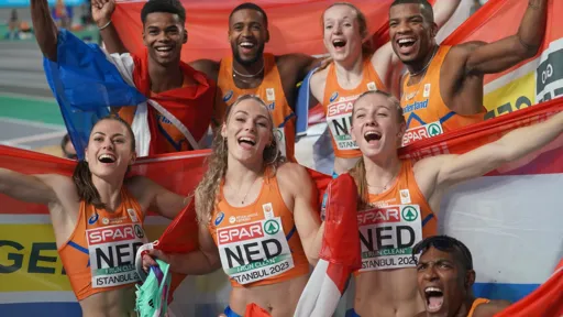 A group of Dutch athletes celebrate together at the European Athletics Indoor Championships in Istanbul 2023. They are wearing orange uniforms with 'NED' on their bibs, holding Dutch flags, and smiling joyfully. Some have their arms raised in victory, while others have their arms around each other. The background shows an indoor track stadium with spectators.