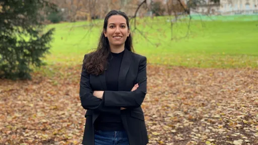 This is a picture of Grace Zakka in a black suit, standing smiling with her arms folded in the foreground with a park in the background.