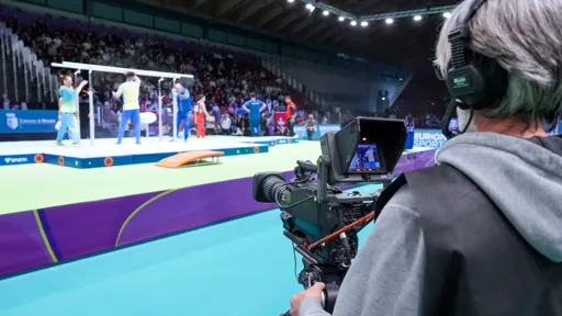 A cameraman records a gymnastics event inside a sports arena, focusing on a group of male gymnasts near the parallel bars. The camera monitor displays the live footage, while the arena is filled with spectators in the background.