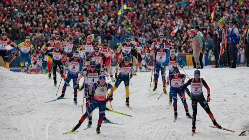 Start of the Women's Mass Start biathlon race on February 23, 2025. A large group of female biathletes, wearing numbered bibs and carrying rifles on their backs, ski forward from the starting area. The background features a packed grandstand with cheering fans, many waving flags.