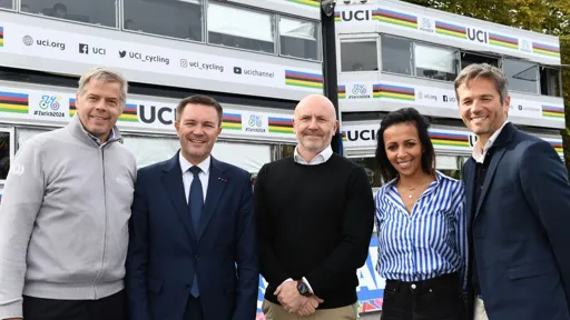 A group of five people stand smiling in front of two double-floor mobile offices decorated with UCI (Union Cycliste Internationale) branding and rainbow stripes. The mobile offices display hashtags and social media handles related to #Zurich2024. Trees are visible in the background.